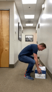 A man demonstrating proper body mechanics as he bends down to pick up a box in a hallway.