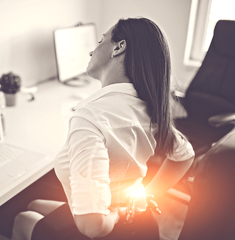 A woman sitting at a desk with her back pain. - Farnsworth Orthopedic